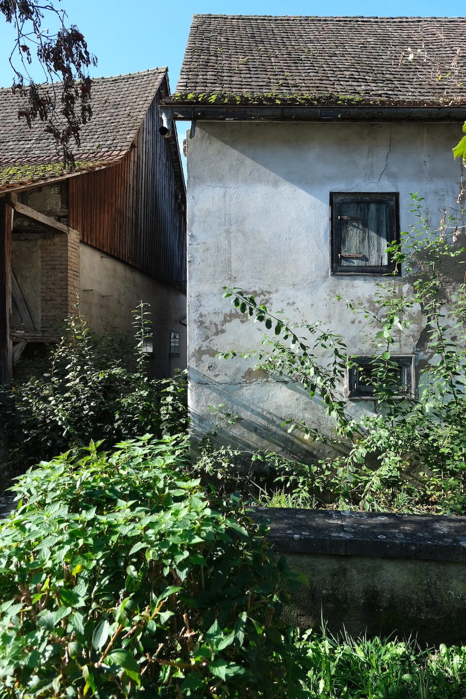 Altes verputztes Gebäude mit verblichenen Fensterläden aus Holz steht neben einer alten Scheune. Im Vordergrund grüne Büsche.